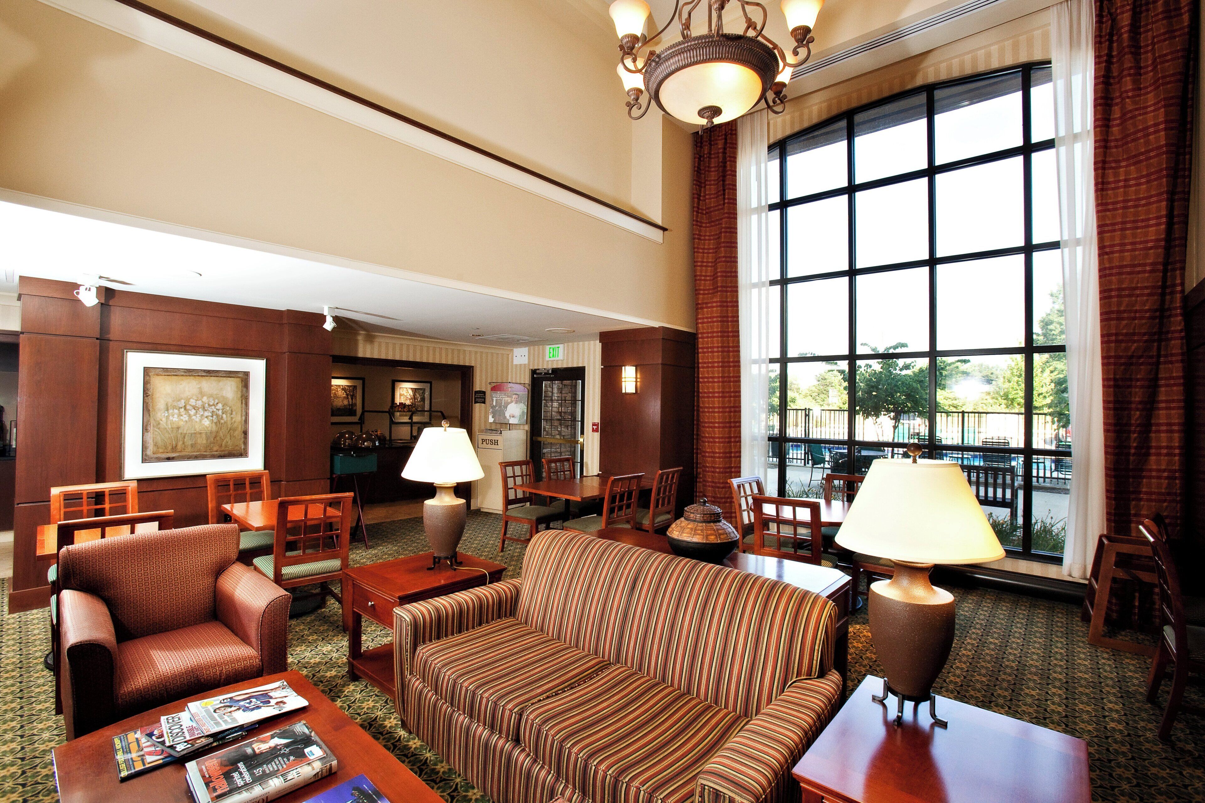 A hotel lobby with a sofa, chairs, a table, a lamp, and a large window offering a view.