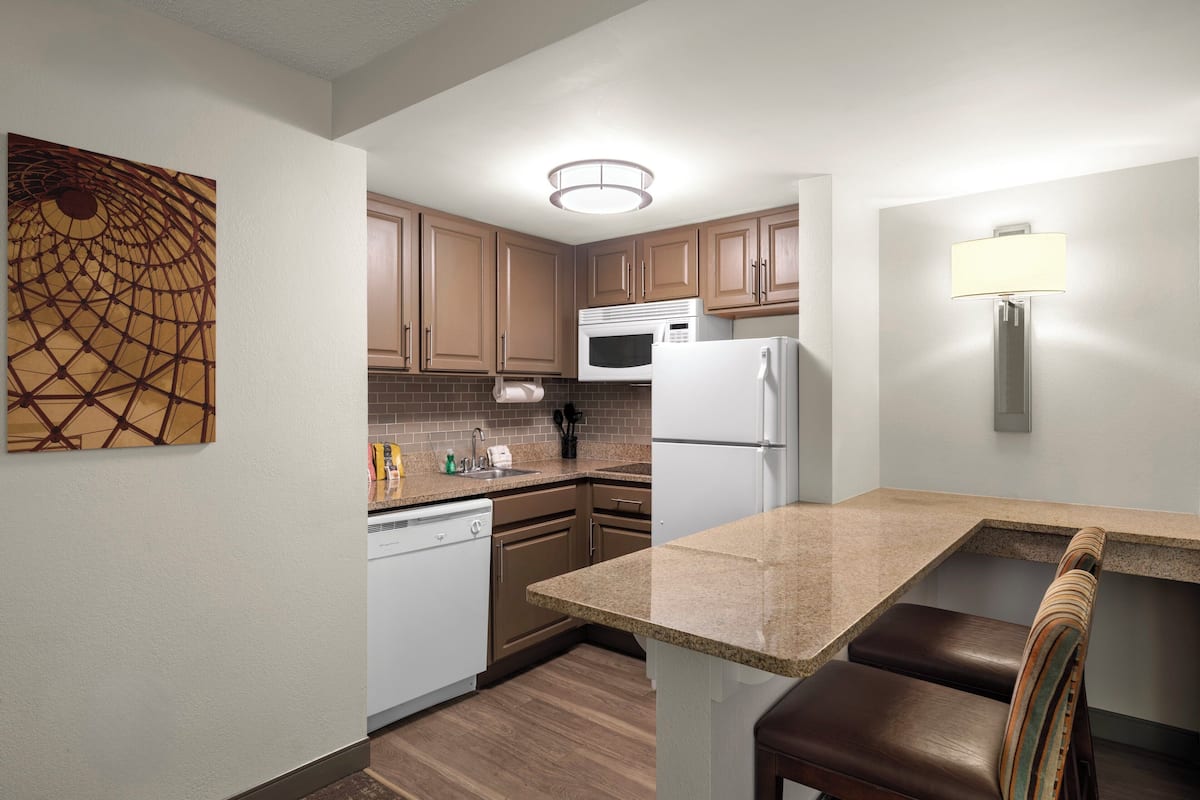 Cocina compacta con armarios de madera, microondas, refrigerador, lavavajillas, lavamanos y una banca.