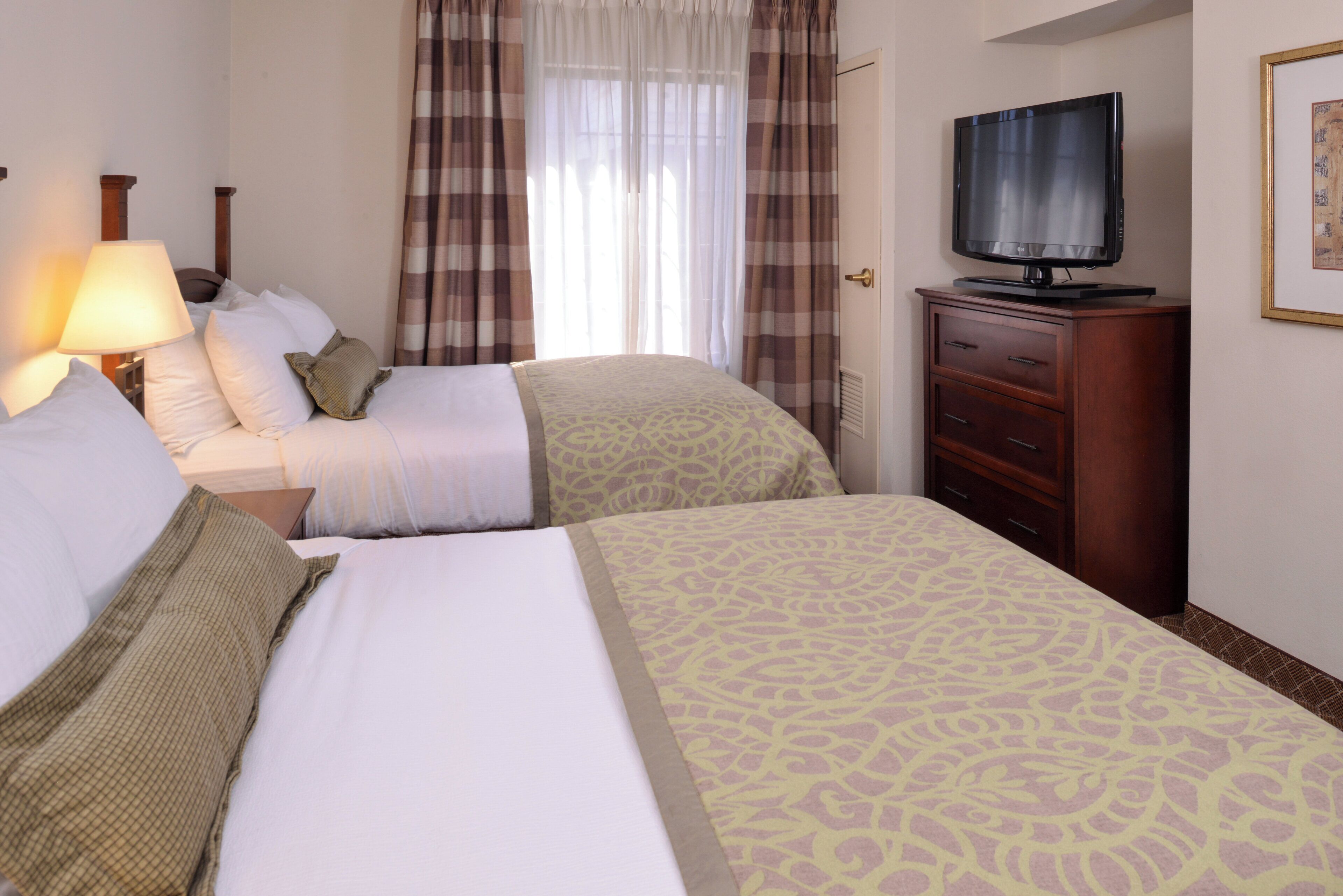 A hotel room with two beds, a television, a dresser, and a window with curtains.