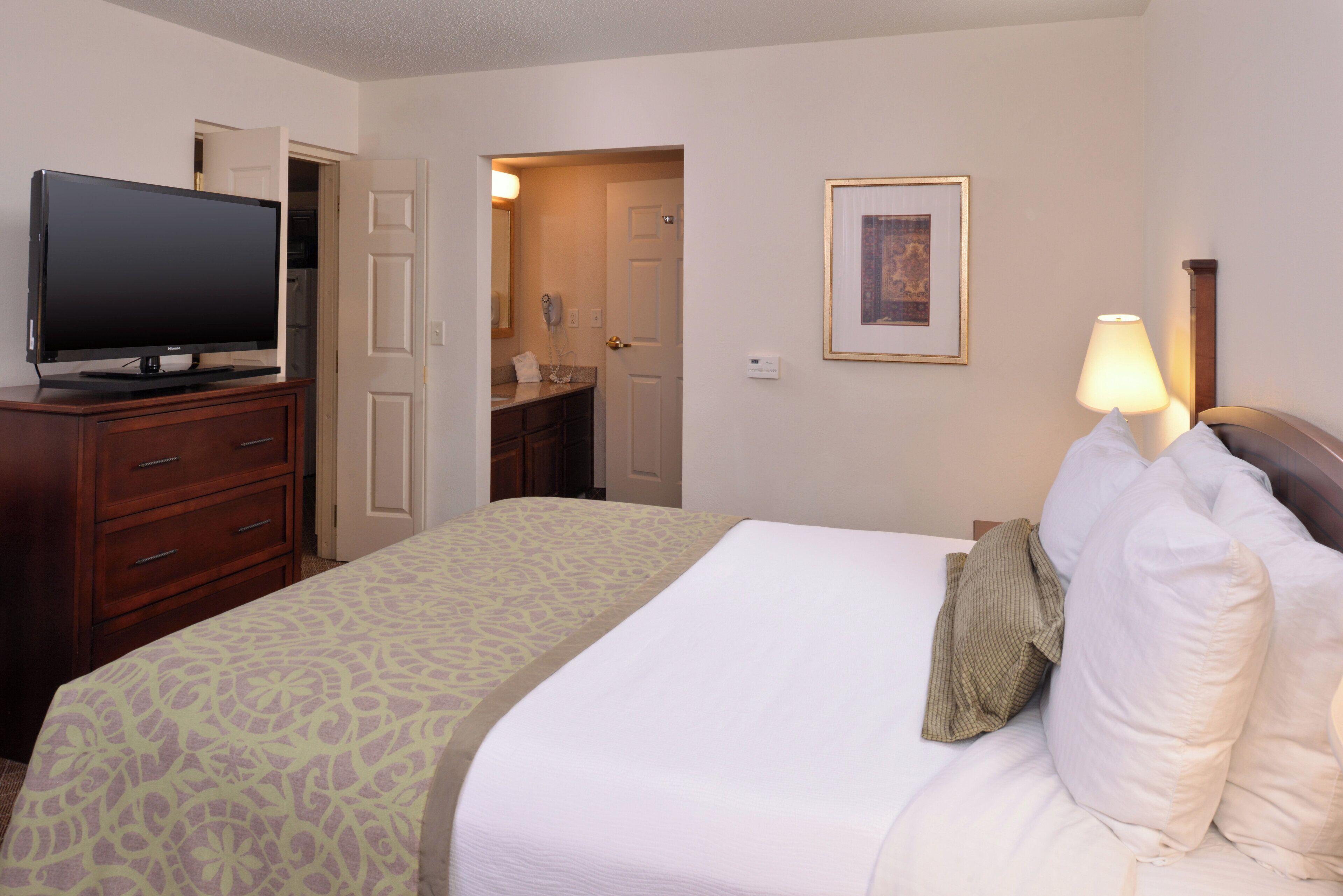 Habitación de hotel con cama, televisor sobre una cómoda, baño visible a través de una puerta abierta y un cuadro en la pared.