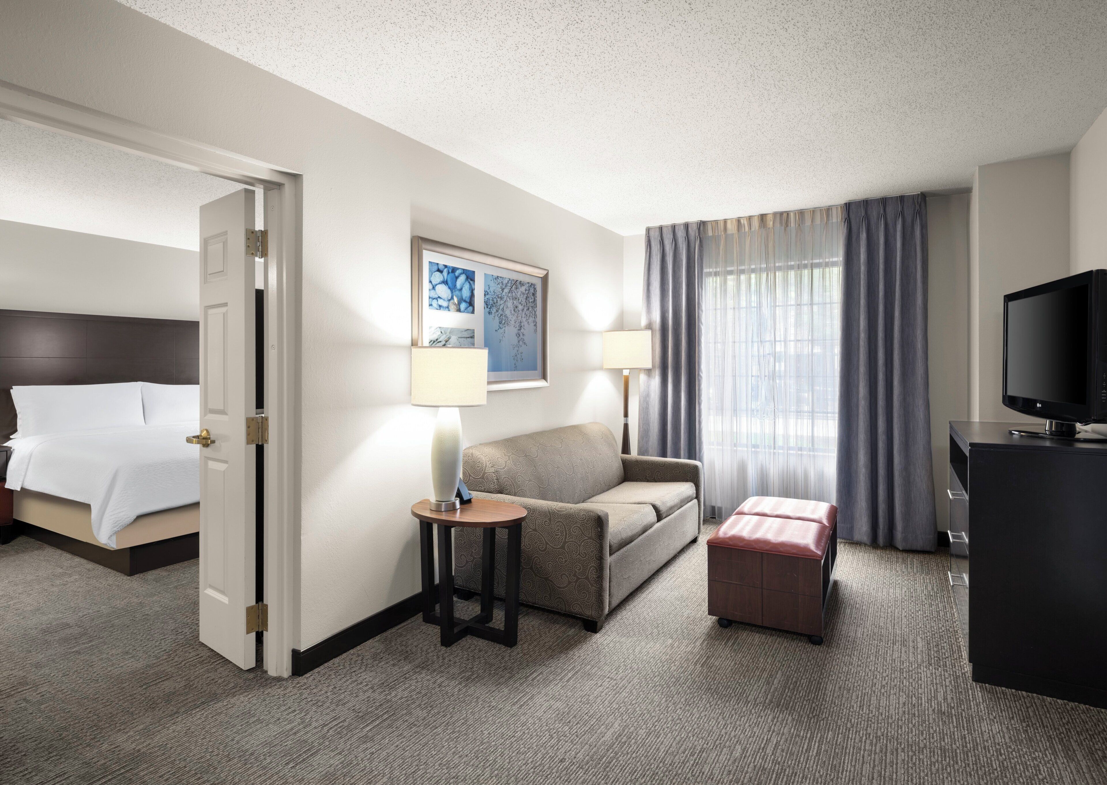 A hotel room with a bed, a sofa, a desk with a television, and a window with curtains.