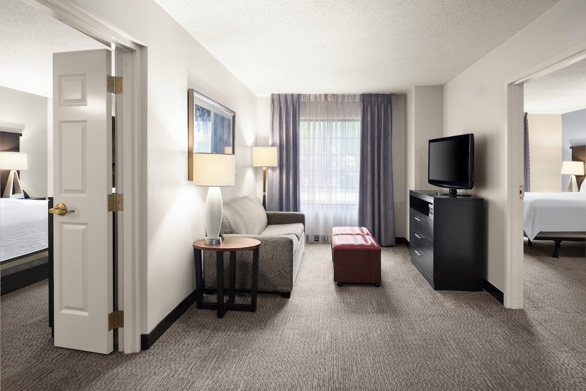 Habitación de hotel con cama, televisión, escritorio con lámpara, silla, mesita y ventana con cortinas.
