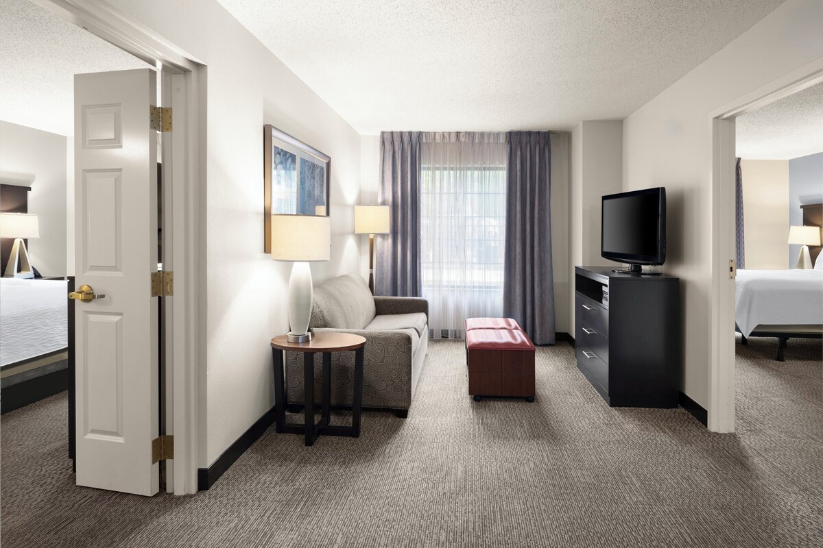 A hotel room with a bed, a desk with a television, a sofa, a small table with a lamp, and a window with curtains.