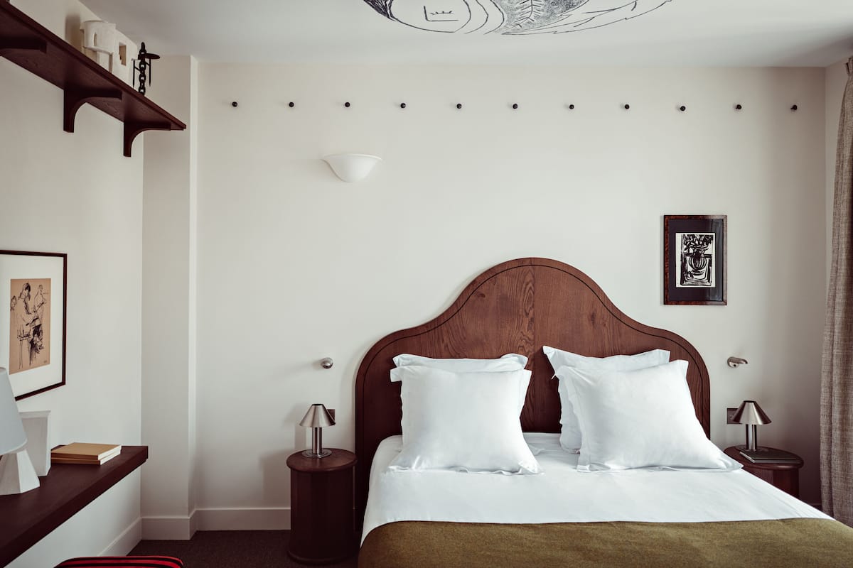A hotel room with a wooden headboard, white bedding, a bedside table with a lamp, a framed picture on the wall, and a small round table with a lamp.