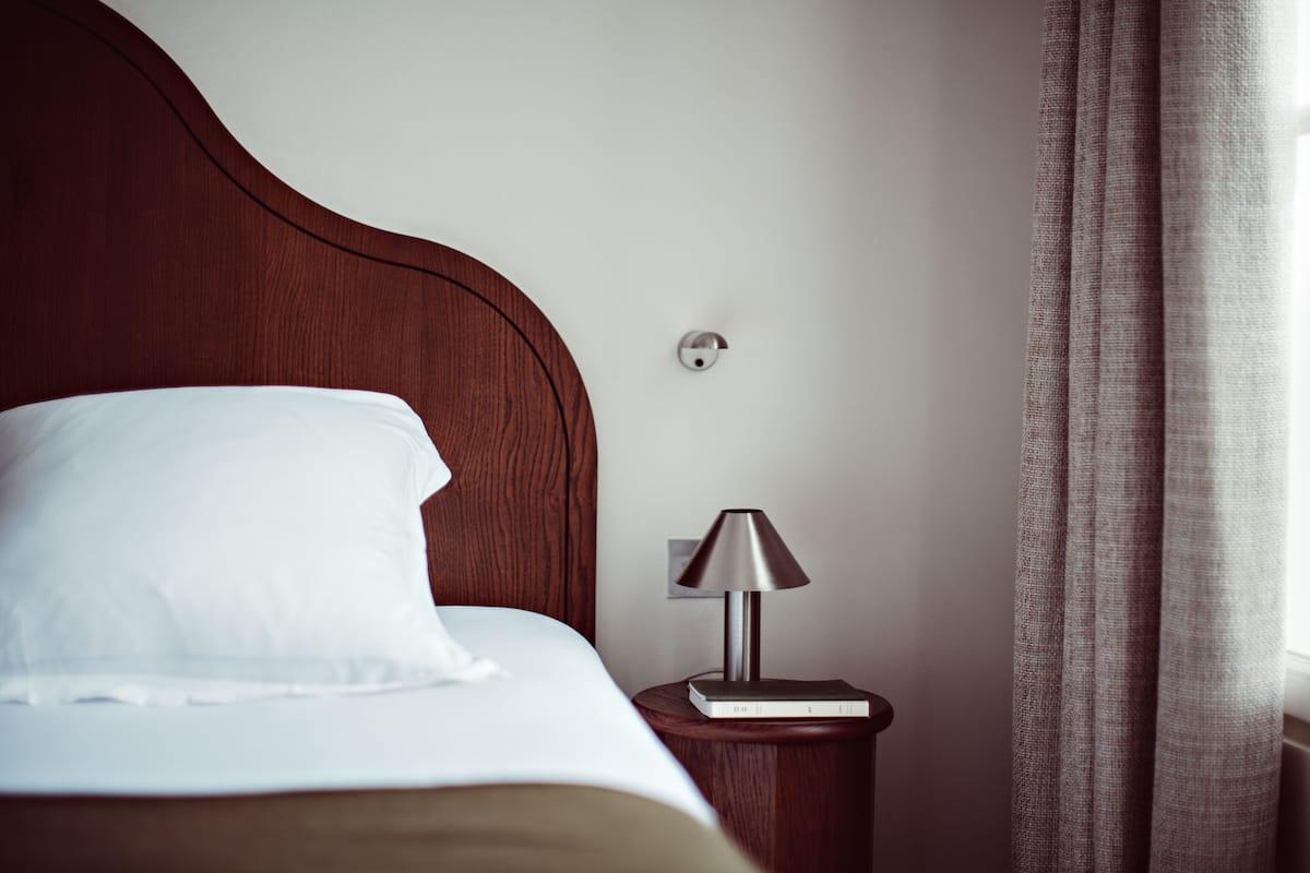 A neatly made bed with a white pillow and a wooden headboard. There is a small bedside table with a lamp and a book.