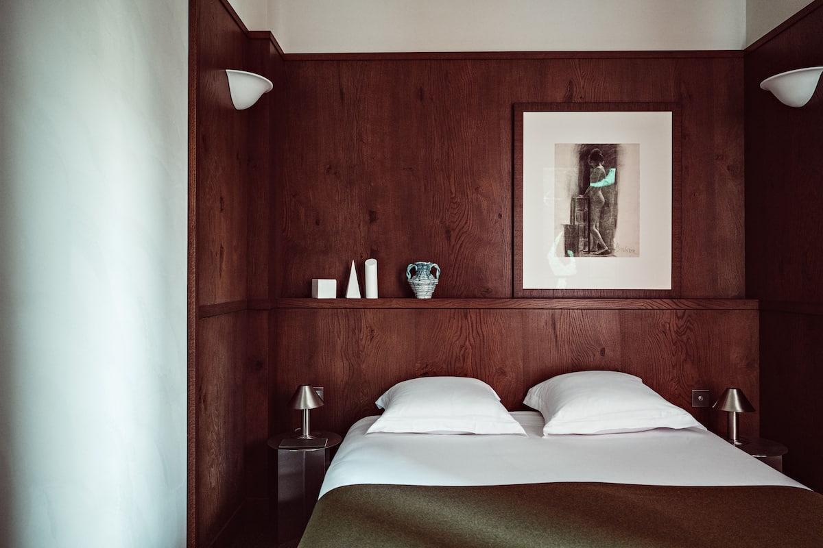 A hotel room with a wooden headboard, a framed artwork, two bedside tables with lamps, and a bed with white linens.