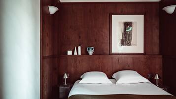 Habitación de hotel con cabecera de madera, una obra de arte enmarcada, dos mesitas de noche con lámparas y una cama con ropa blanca.