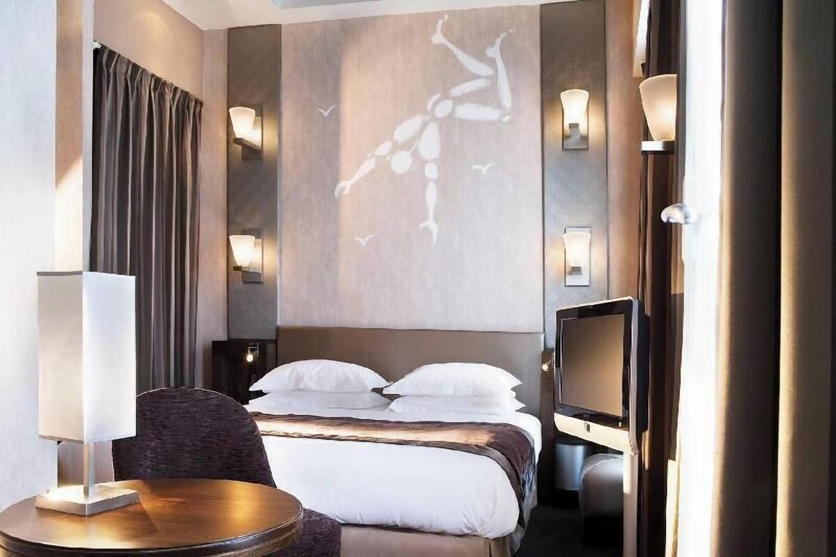 A hotel room with a bed, a TV, a desk, and a wall-mounted light fixture.