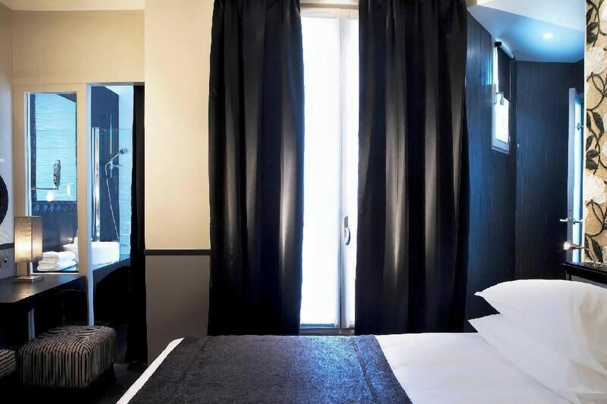 A hotel room with a bed, a desk with a lamp, a mirror, and a window with curtains.