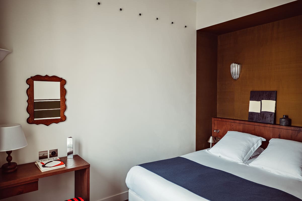 A hotel room with a neatly made bed, a wooden nightstand with a lamp, a mirror, and a framed picture.