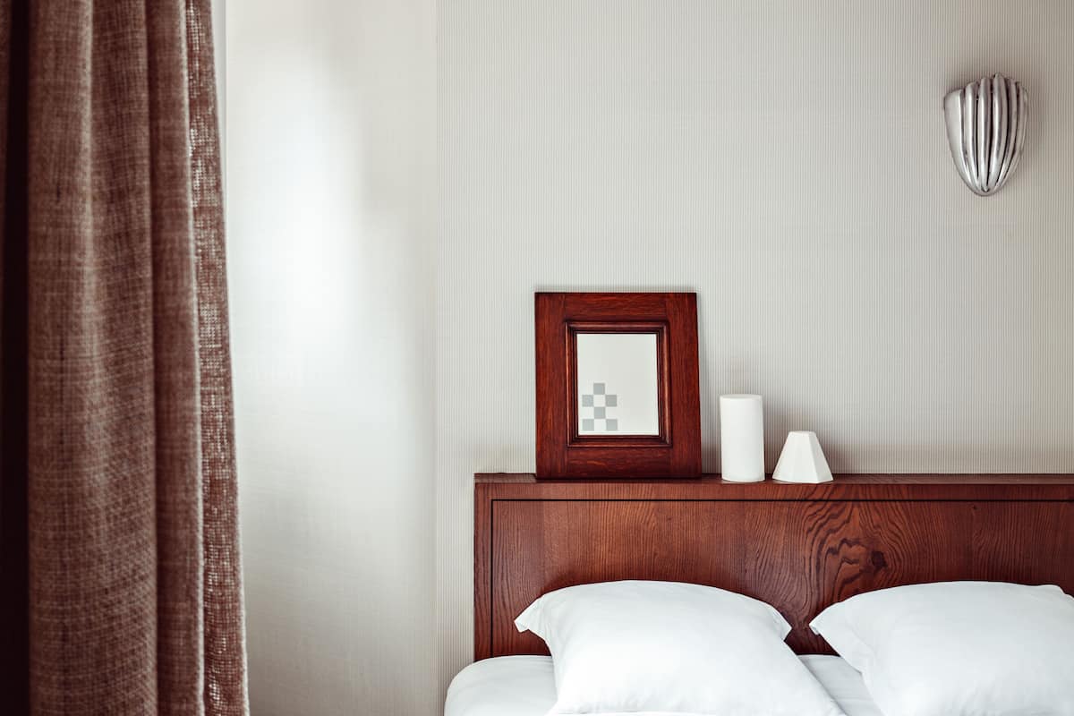 A neatly made bed with white linens, a wooden headboard, a framed picture, a white candle, and a wall-mounted light fixture.
