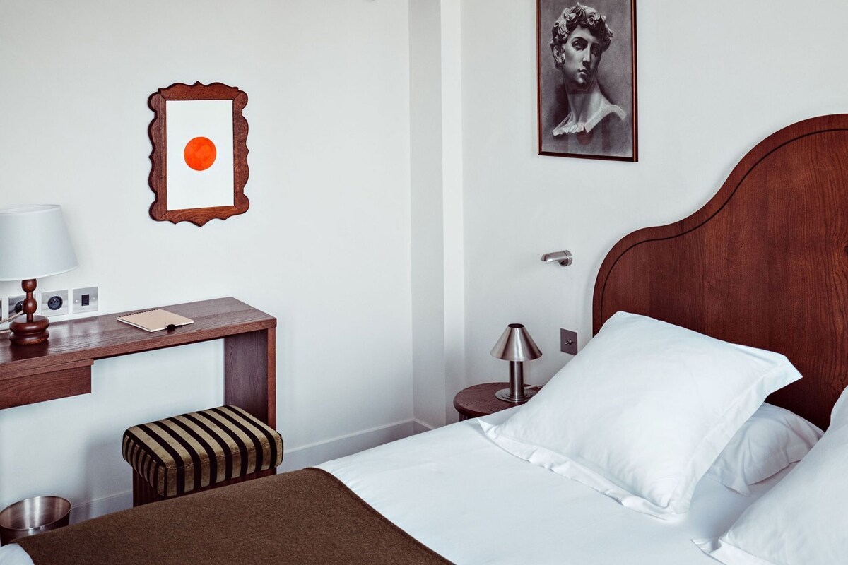 A hotel room with a wooden bed, a desk with a lamp, a small stool, and a framed picture on the wall.