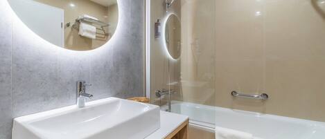 A modern bathroom with a large sink, a bathtub, a glass-enclosed shower, and a round mirror.