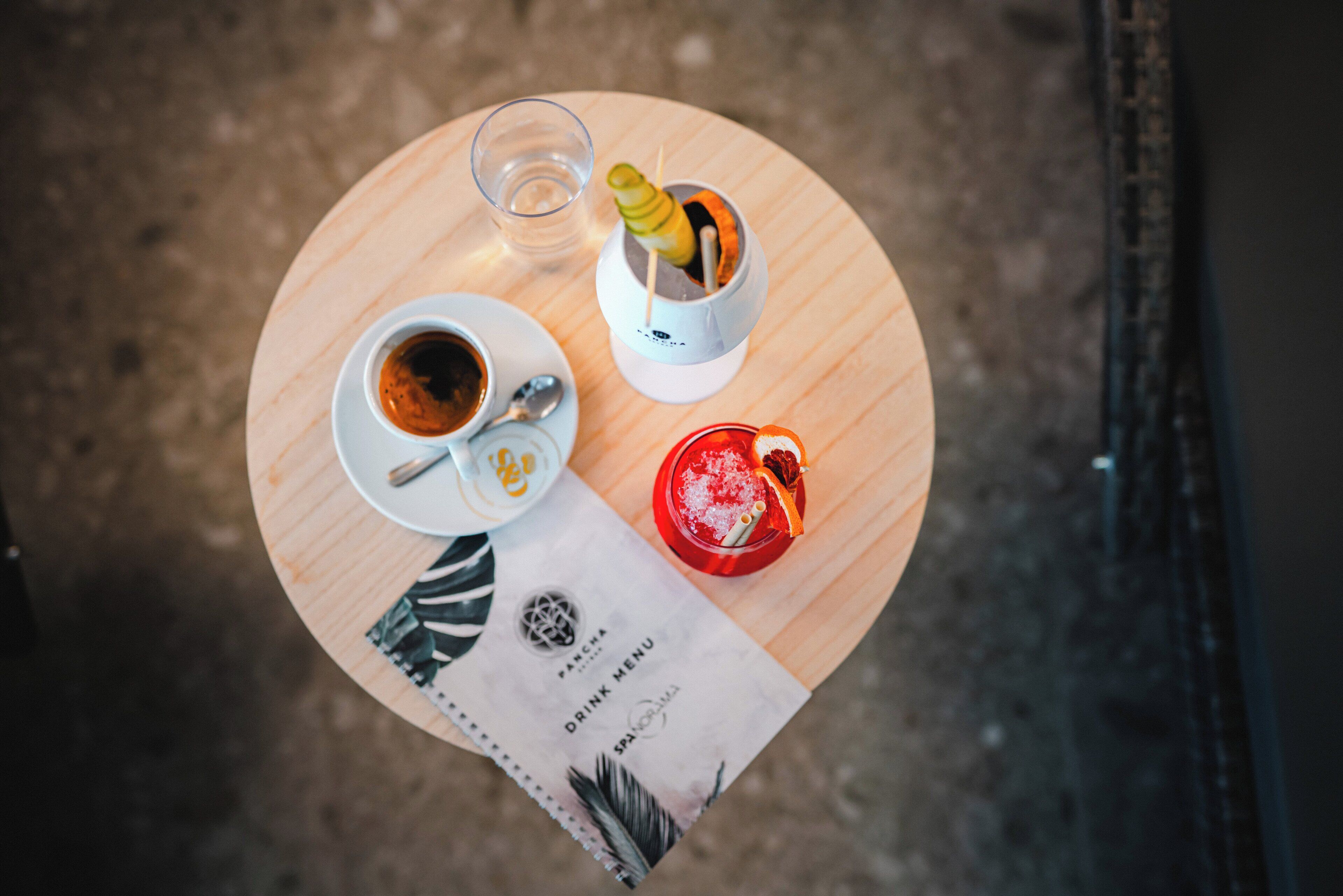 A round table with a menu, a cup of coffee, a glass of red drink, and a small white cup with a yellow item.