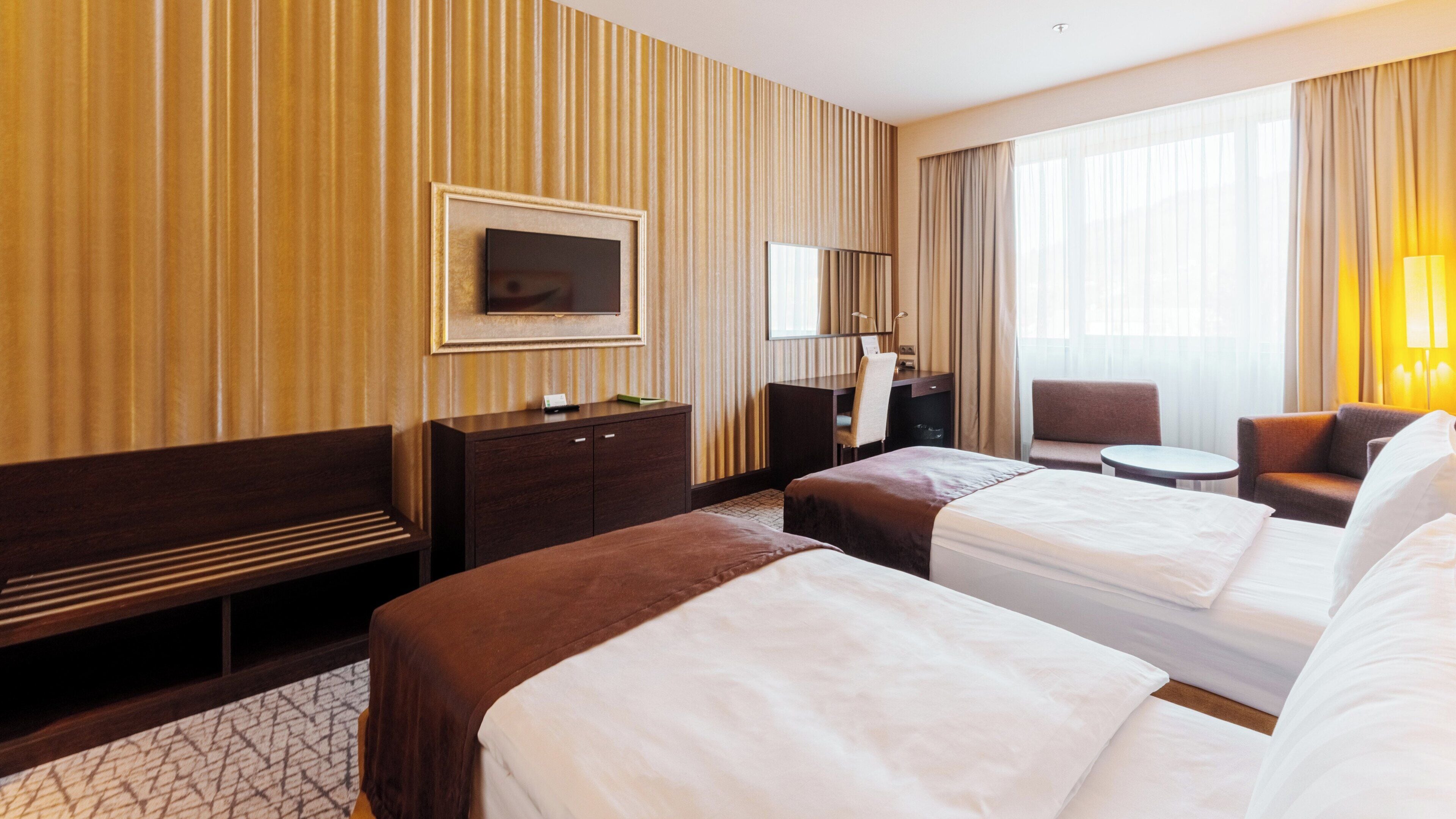 A hotel room with two beds, a desk, a chair, a TV, and a window with curtains.