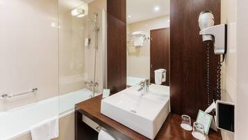 A modern bathroom with a glass shower enclosure, a white bathtub, a wooden vanity with a white sink, and a hairdryer mounted on the wall.
