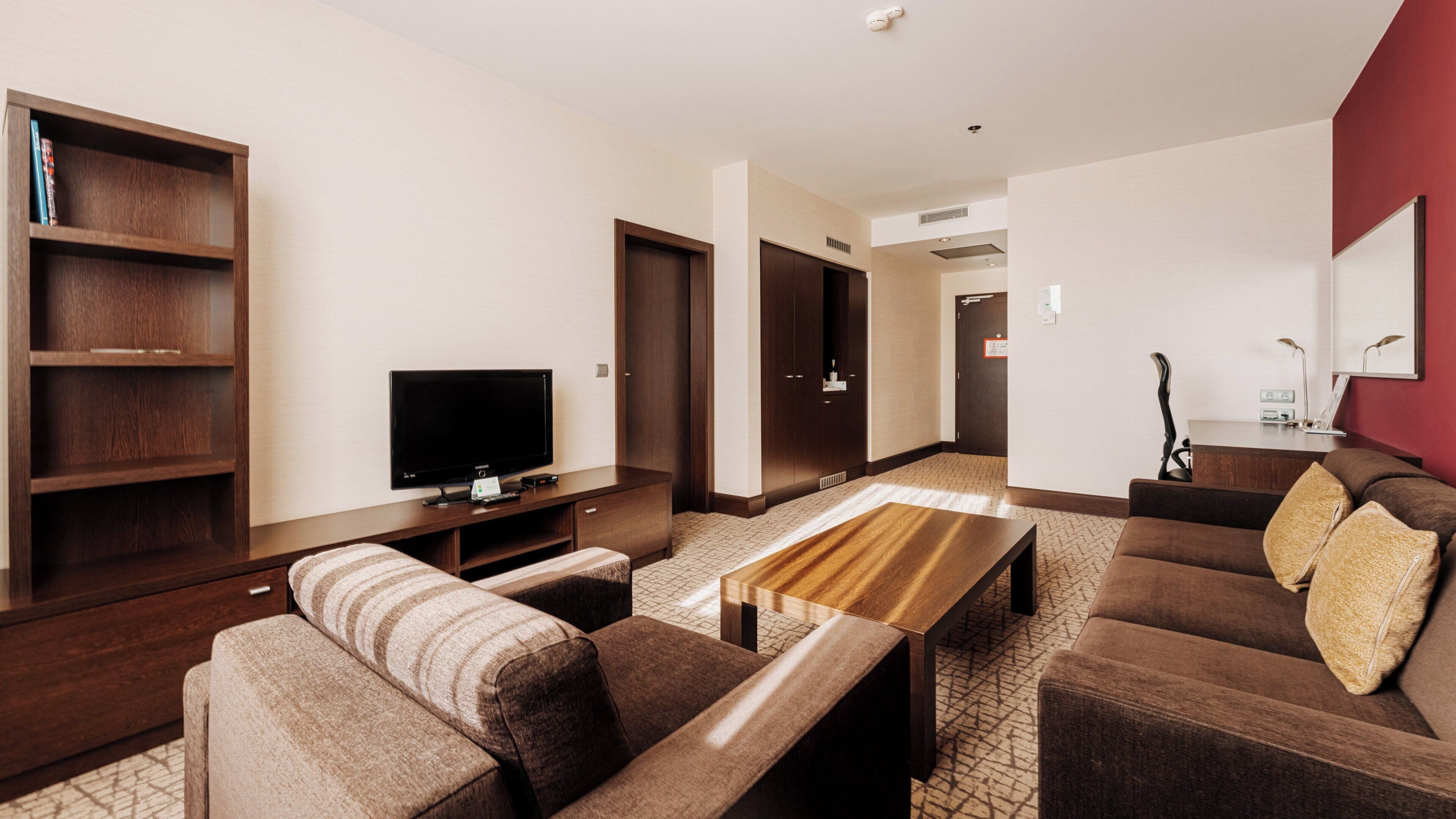 A modern hotel room with a flat-screen TV, a wooden entertainment unit, a sofa, a coffee table, and a red accent wall.