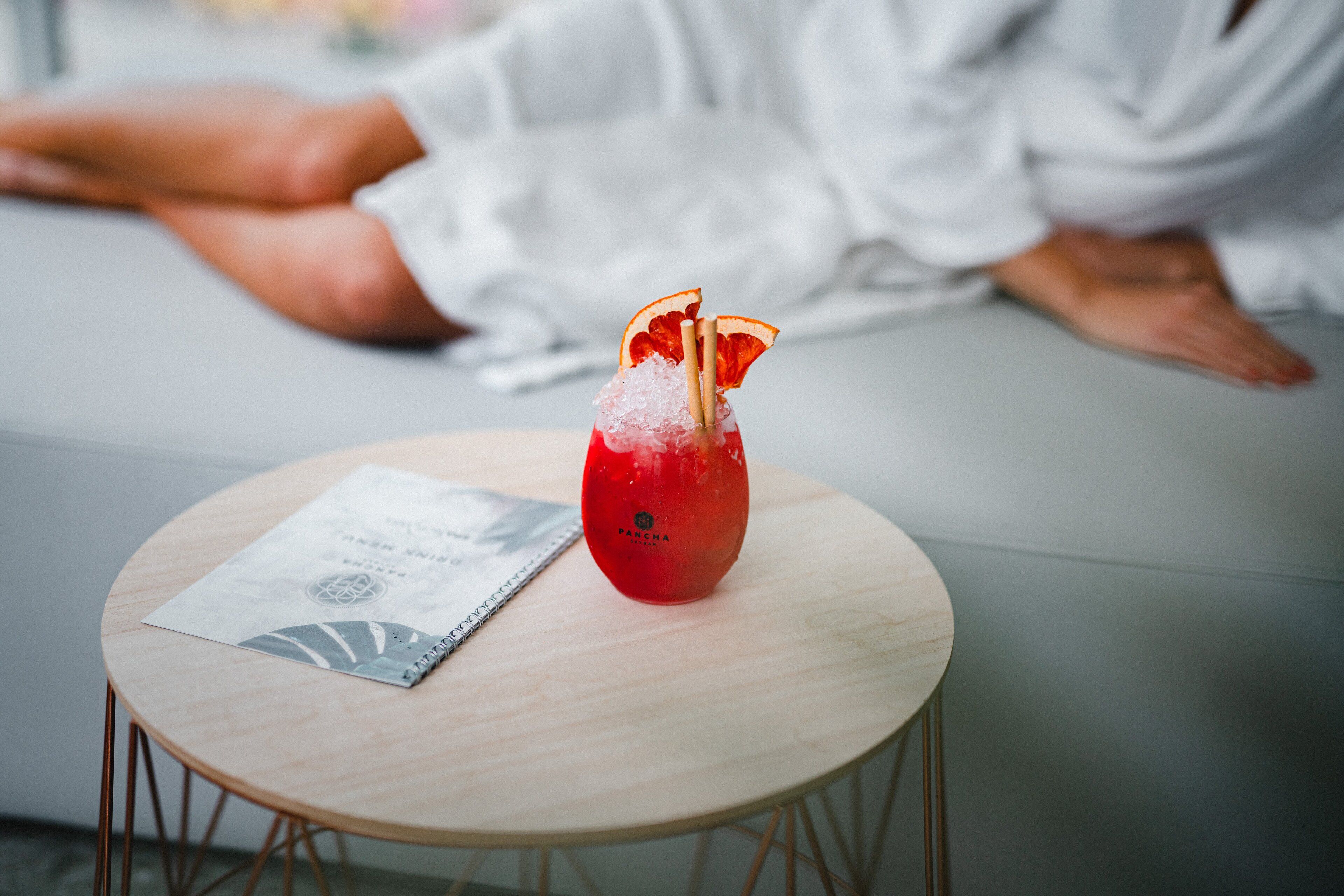 A round wooden table with a red drink garnished with an orange slice and a straw, and a menu or pamphlet on it.