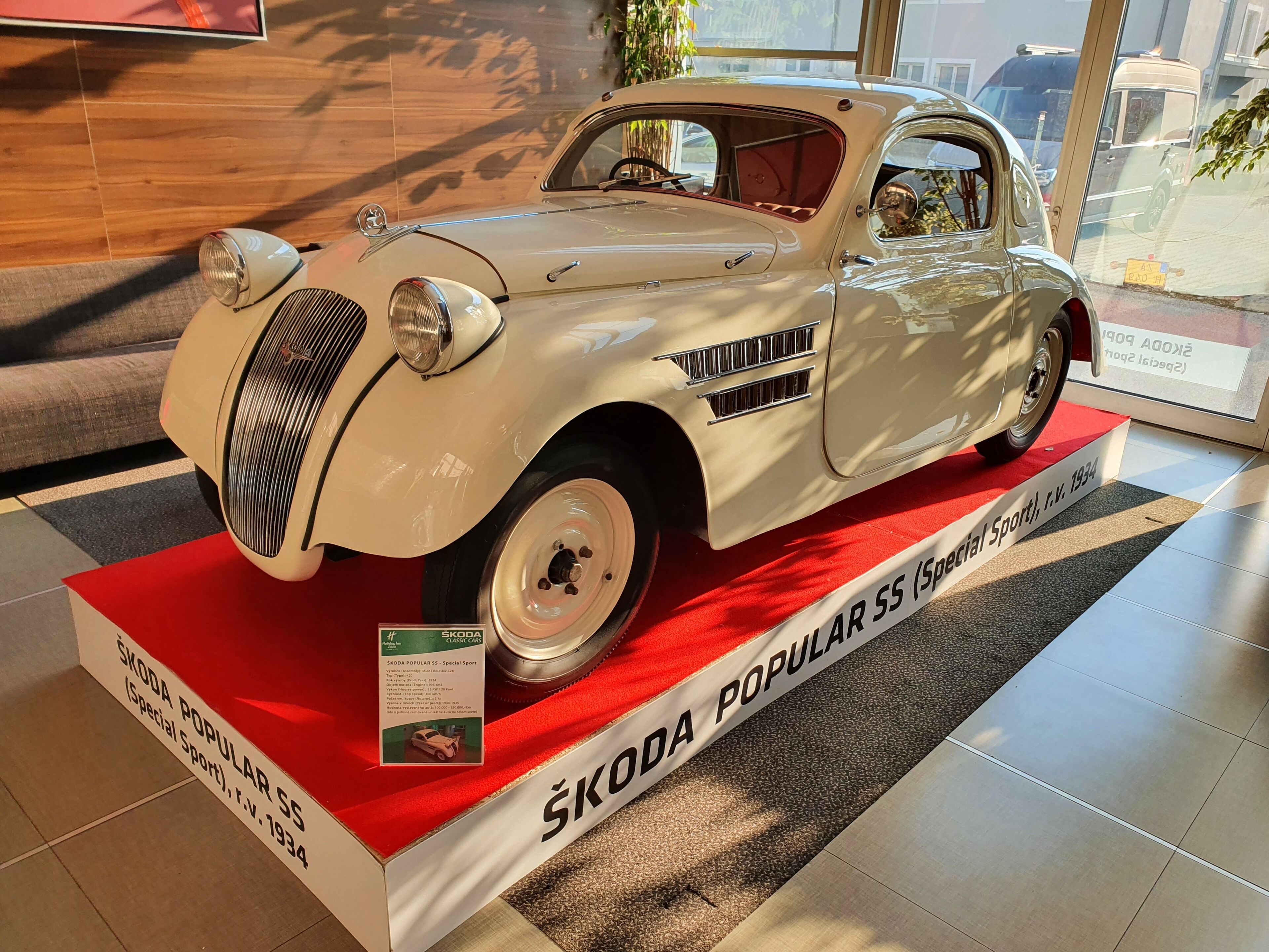 Un coche clásico ŠKODA POPULAR SS expuesto sobre una alfombra roja y un cartel informativo.