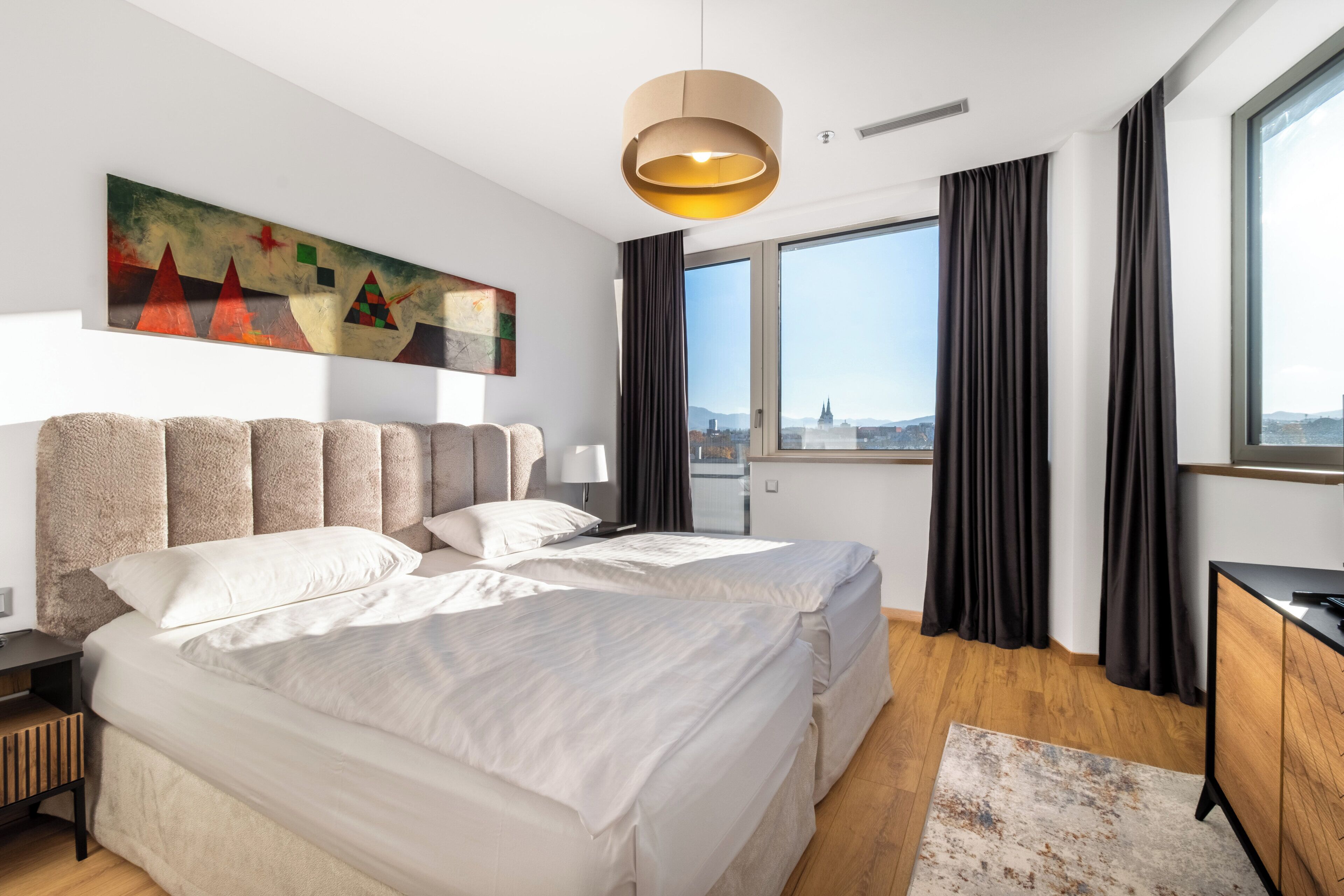 Ett modernt hotellrum med en stor säng, ett skrivbord och utsikt över staden genom stora fönster.