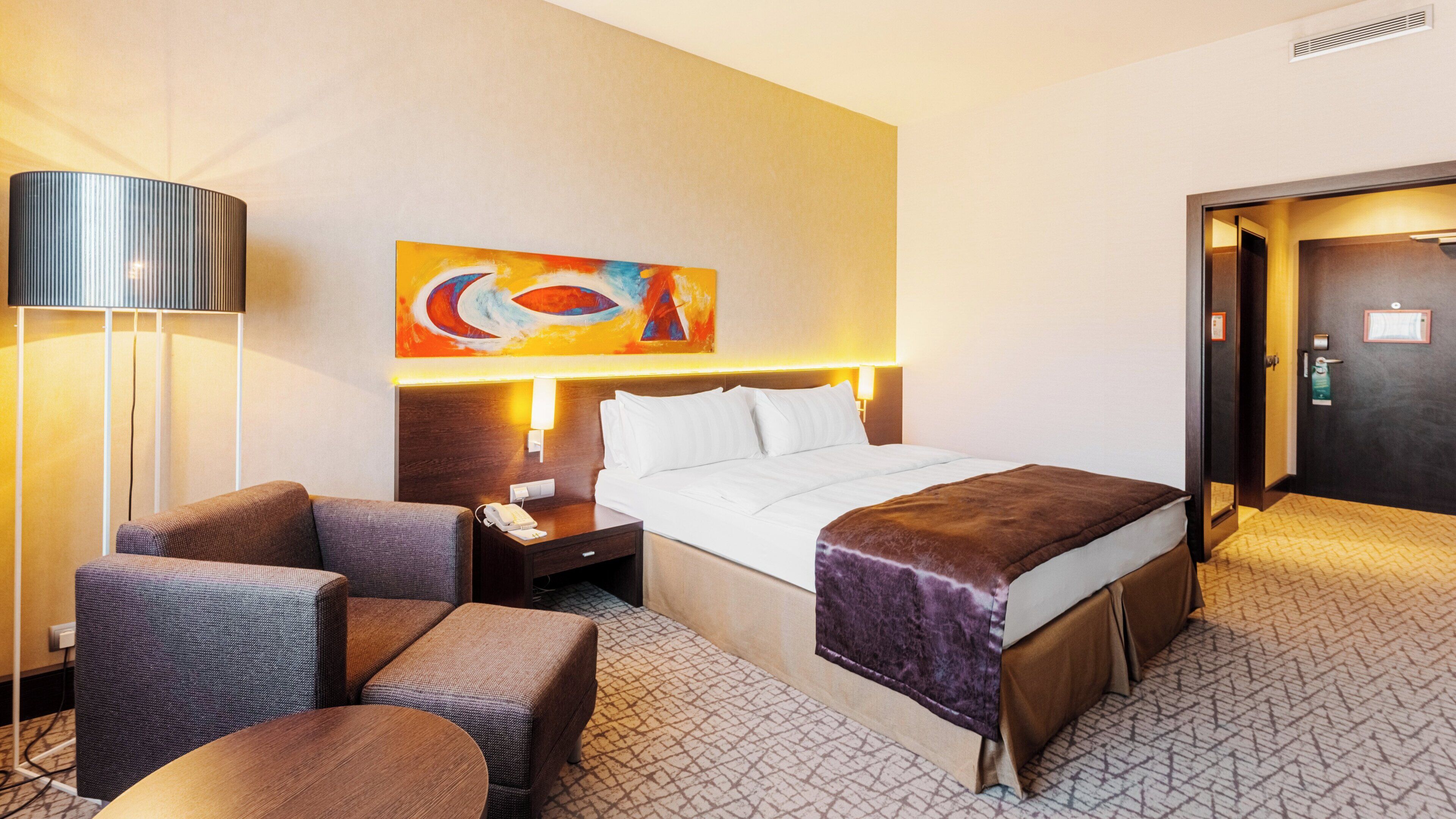 A hotel room with a bed, two armchairs, a nightstand, a lamp, and a painting on the wall.