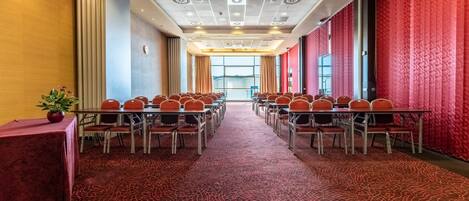 Un salón de conferencias con mesas y sillas, alfombra estampada y un muro con cortinas rojas.