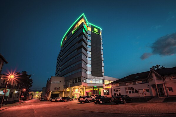 A multi-story building with a Holiday Inn sign, a parking area with several cars, and a street with street lamps.
