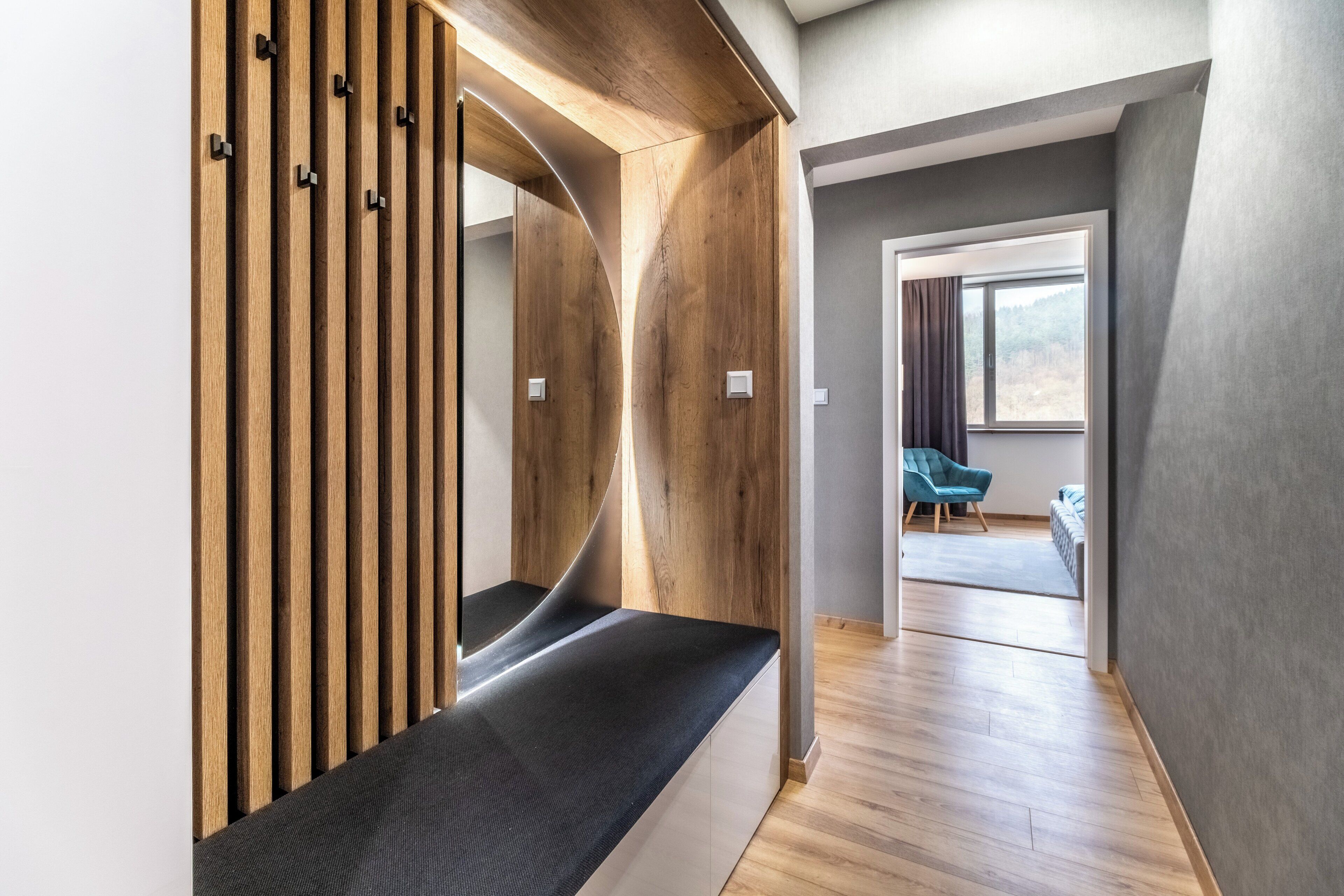 Un intérieur moderne avec un mur à lattes de bois, un banc et une vue sur une pièce où se trouvent une chaise bleue et une fenêtre.