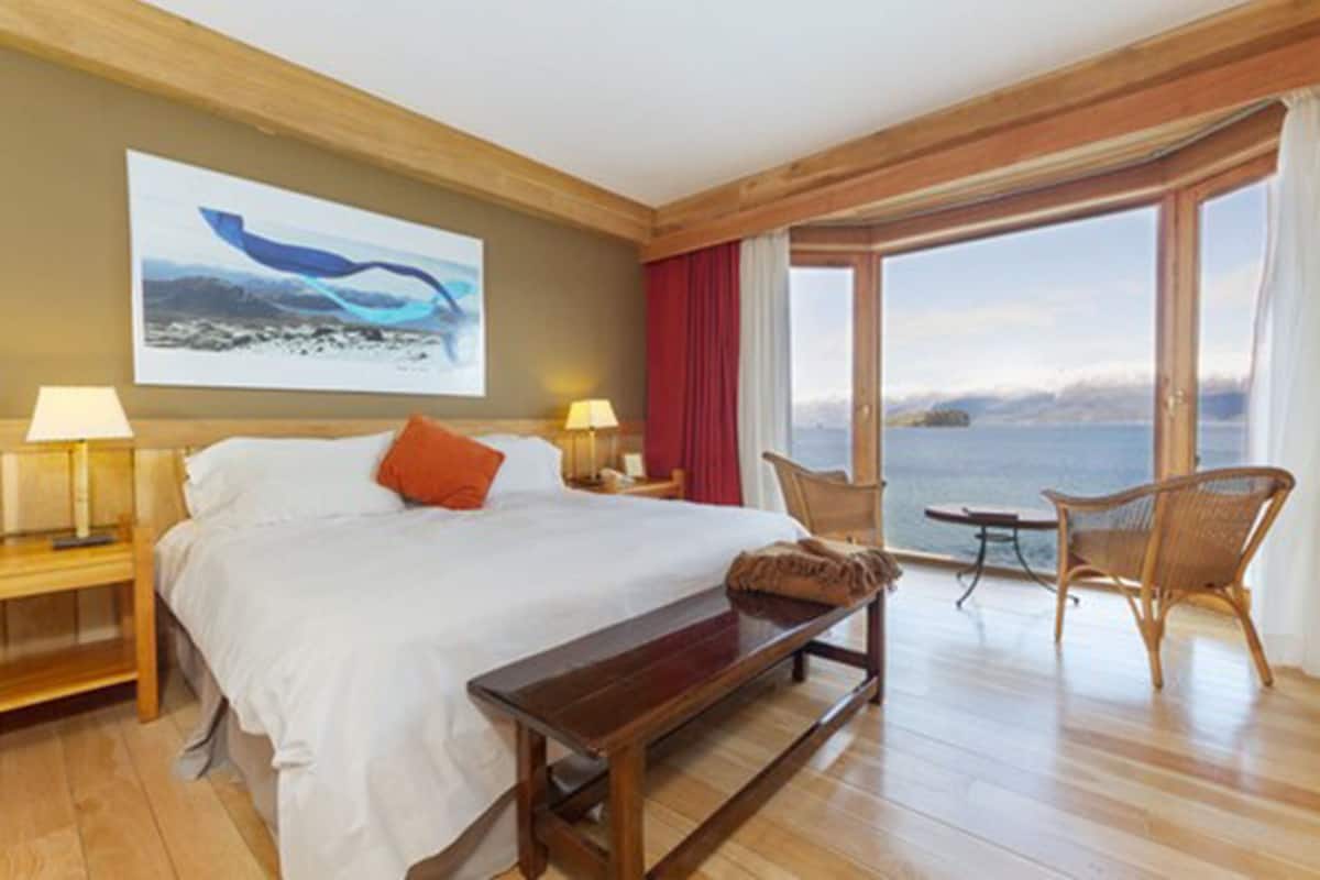 Ett hotellrum med en stor säng, en träbänk, en stol, ett bord och utsikt över havet.