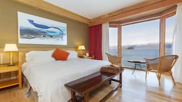 A hotel room with a large bed, a wooden bench, a chair, a table, and a view of the sea.