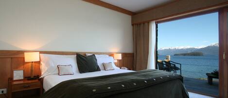 A hotel room with a bed, bedside tables, a lamp, and a view of a lake with mountains in the distance.