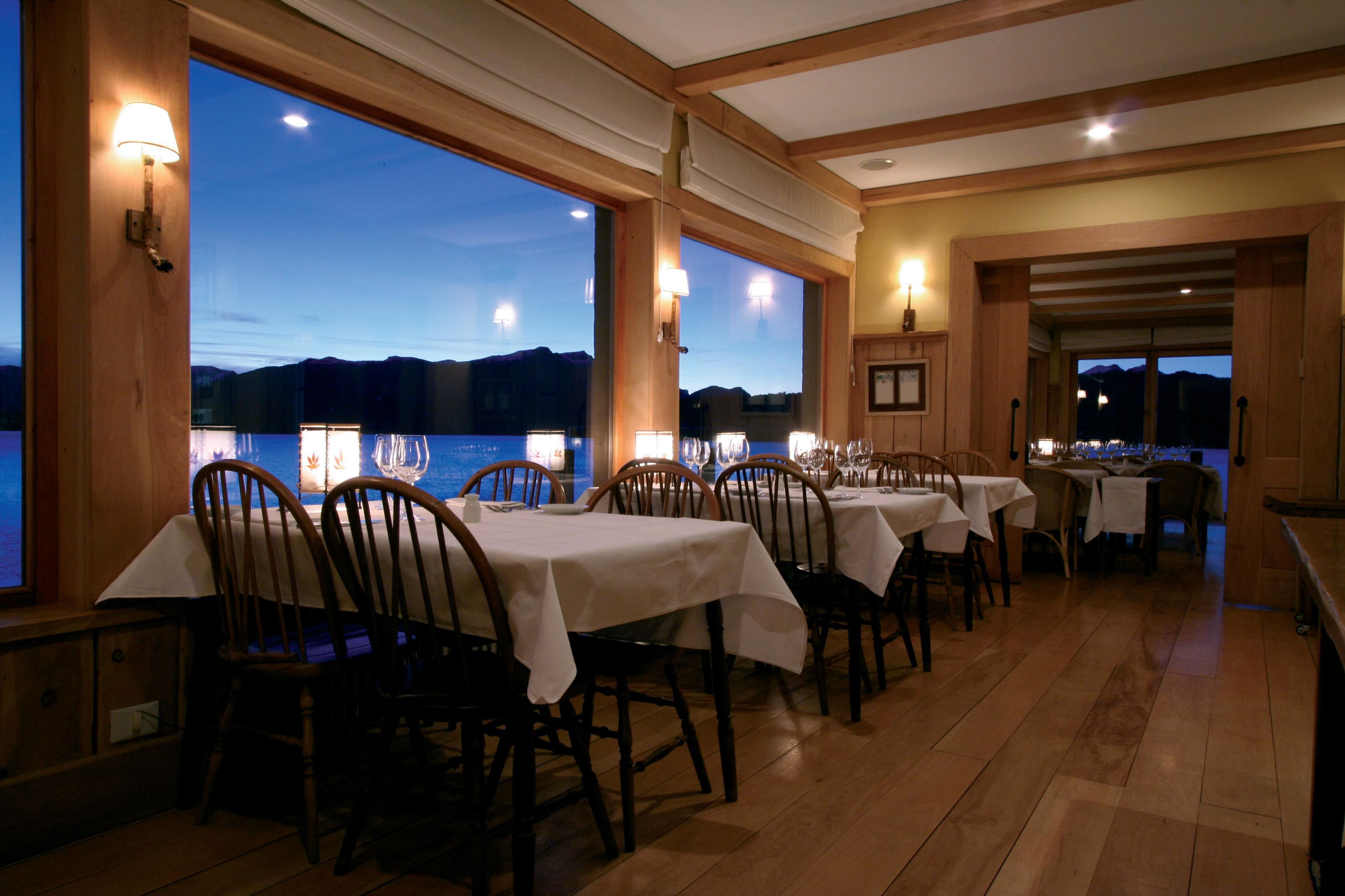 A restaurant with wooden tables and chairs, large windows with a view of the mountains, and warm lighting.