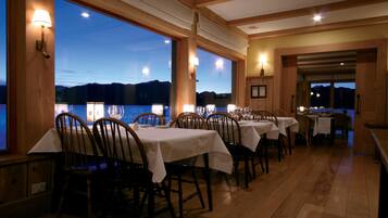 A restaurant with wooden tables and chairs, large windows with a view of the mountains, and warm lighting.