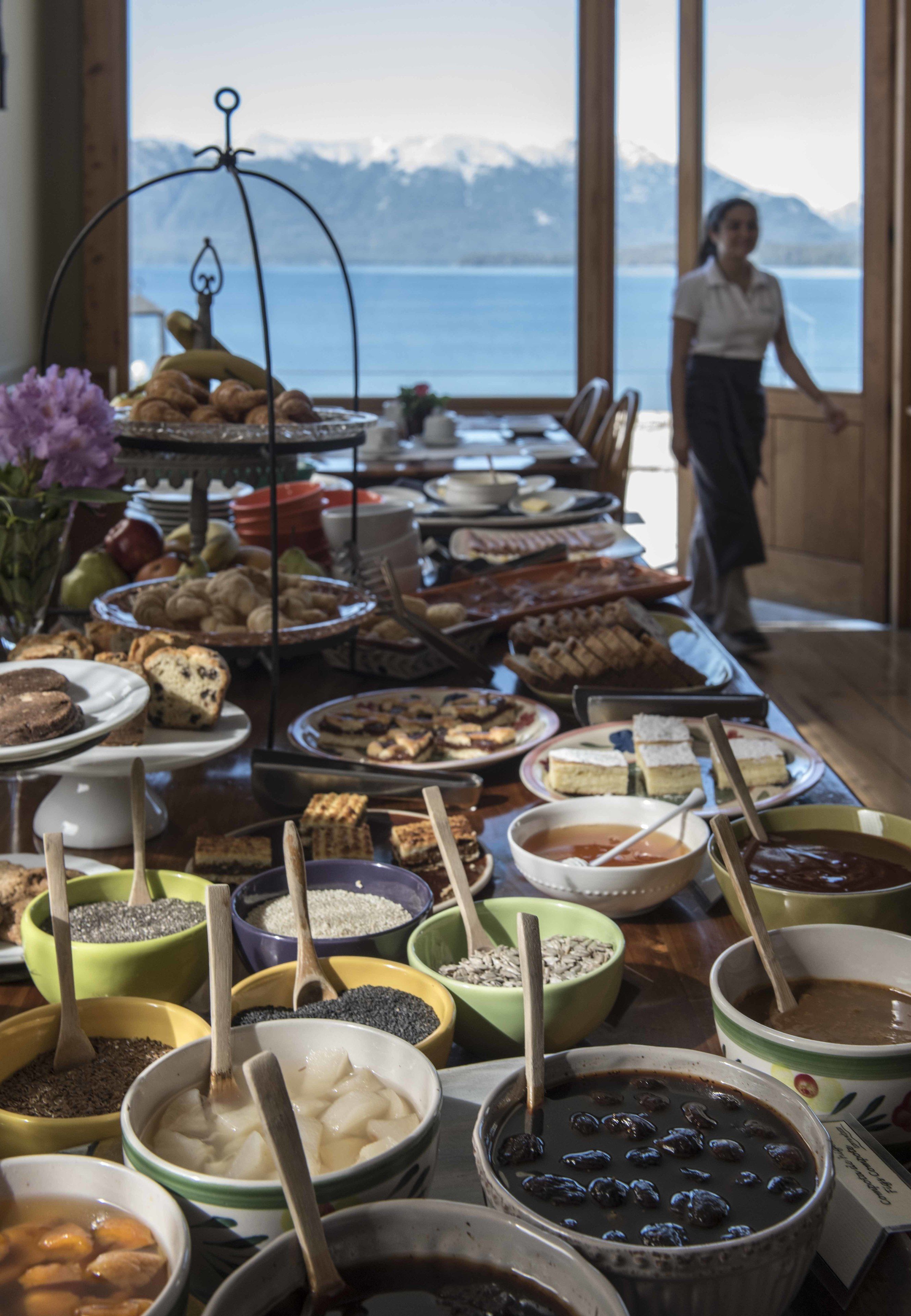 Mesa de buffet com diversos pratos e recipientes, uma pessoa ao fundo e vista para montanhas através de amplas janelas.