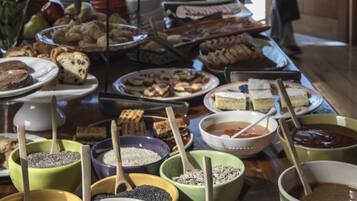 Mesa de buffet com diversos pratos e recipientes, uma pessoa ao fundo e vista para montanhas através de amplas janelas.