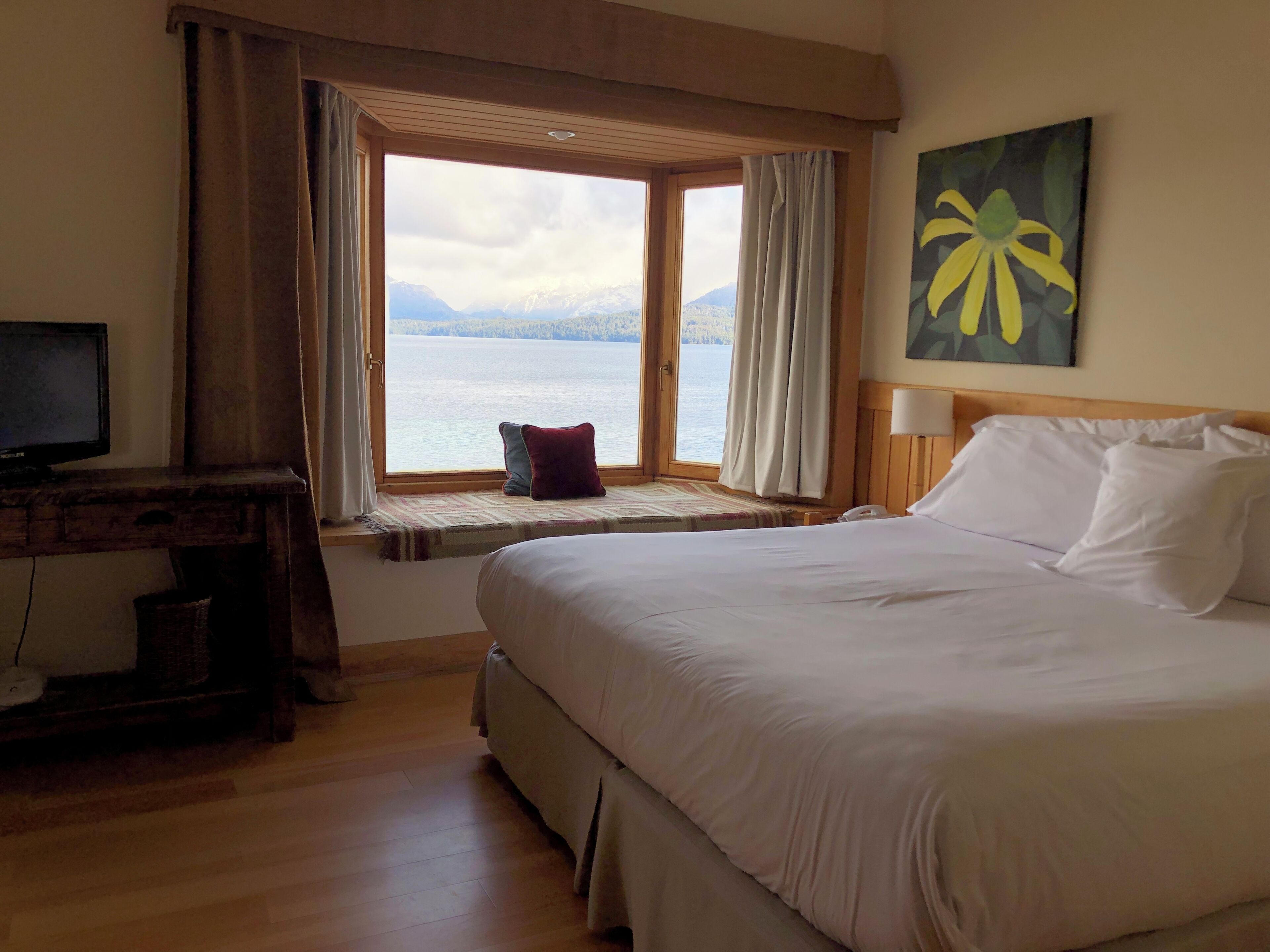 Habitación de hotel con cama, escritorio con televisor, ventana con cortinas y un cuadro en la pared.