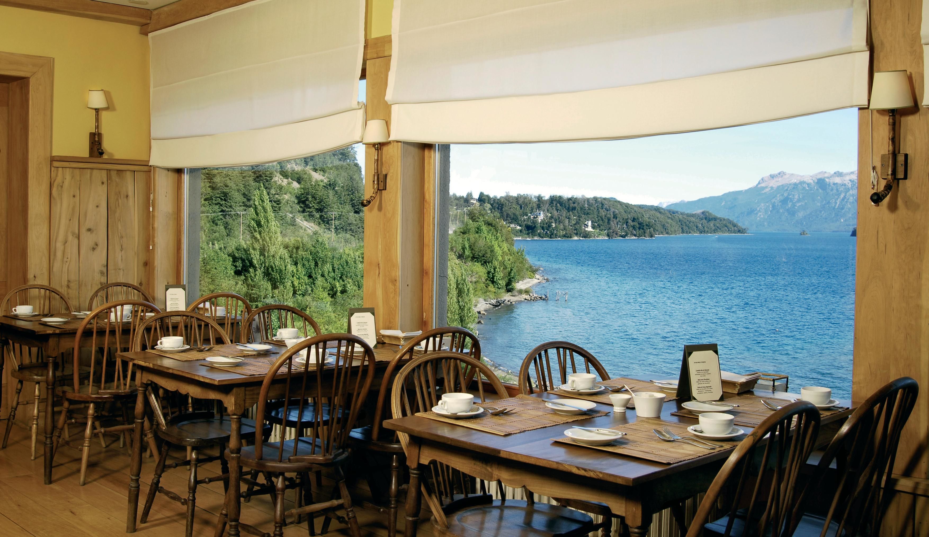 A dining area with wooden tables and chairs, set against a backdrop of a lake and mountains.