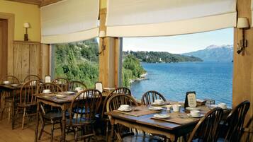 A dining area with wooden tables and chairs, set against a backdrop of a lake and mountains.