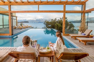 A couple seated at a wooden table by an infinity pool with a view of a lake and mountains.