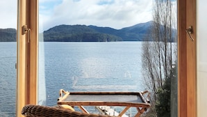 A room with a large window overlooking a lake, a wicker chair, and a small table. - Correntoso Lake & River Hotel (Villa La Angostura)