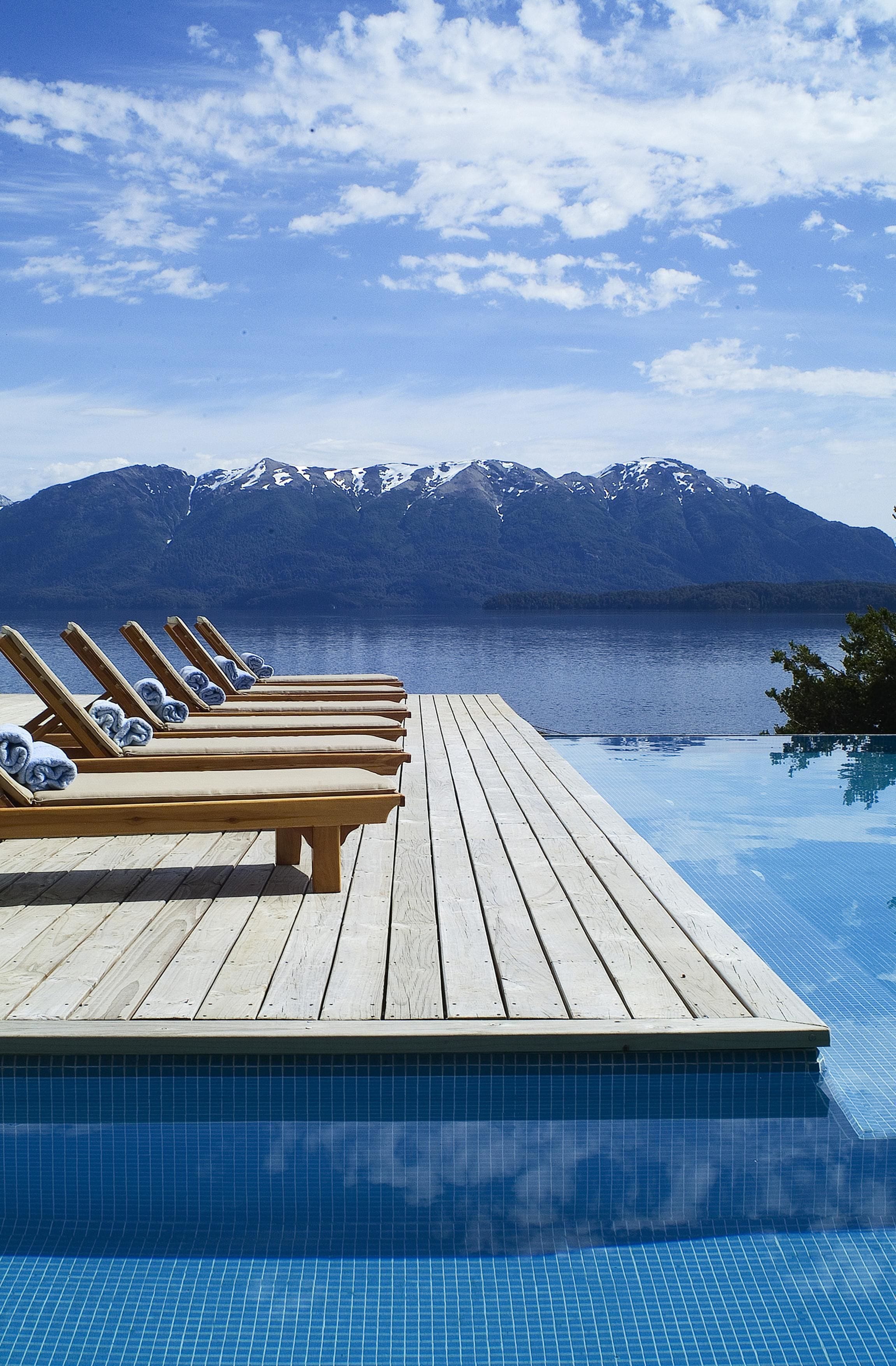 Um deck de madeira com cadeiras de descanso e piscina, com vista para um lago e montanhas ao fundo.