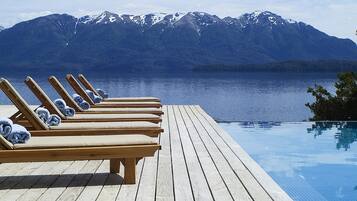 Um deck de madeira com cadeiras de descanso e piscina, com vista para um lago e montanhas ao fundo.