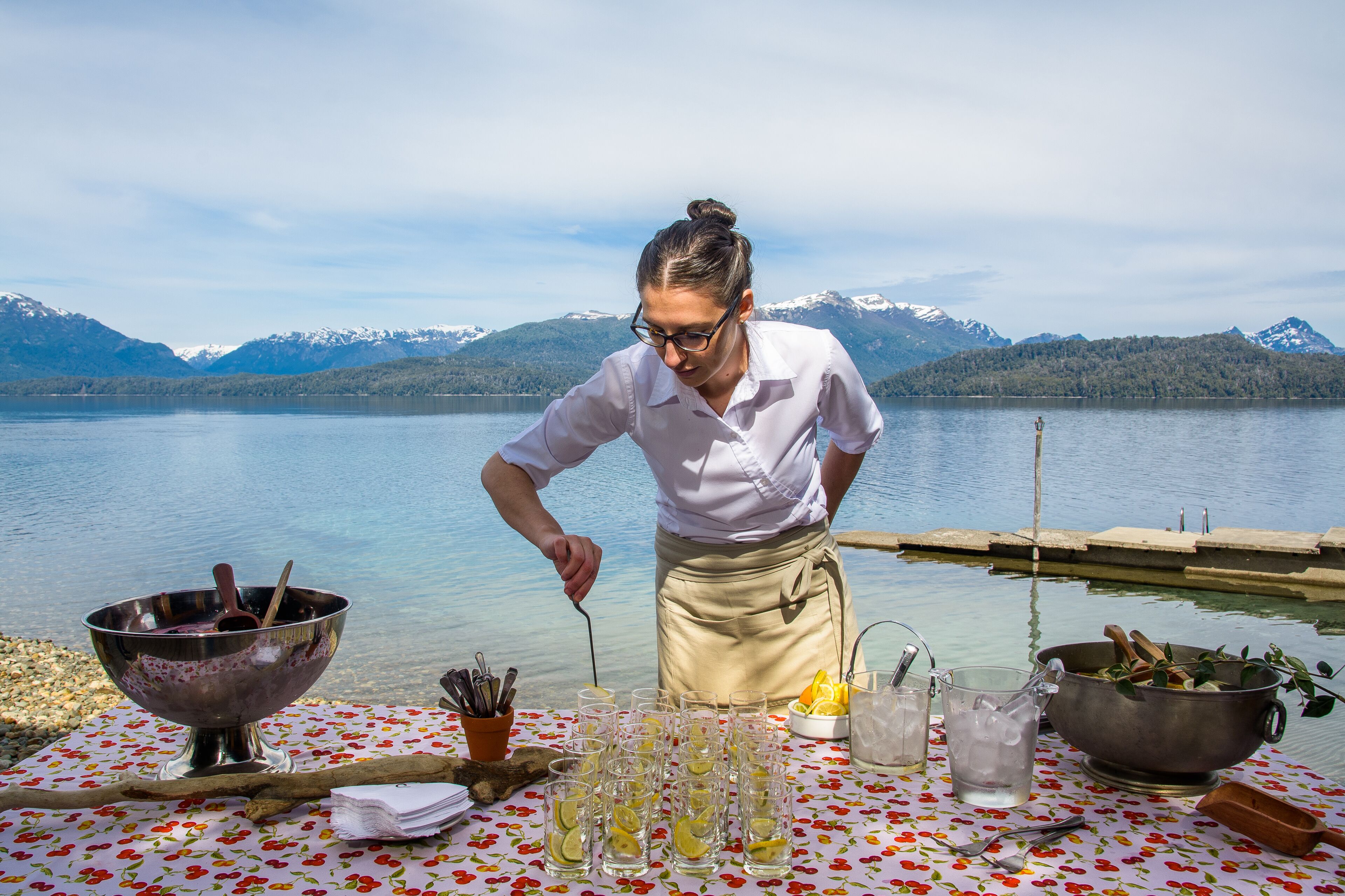 A person preparing drinks at a lakeside table with a scenic mountain backdrop.