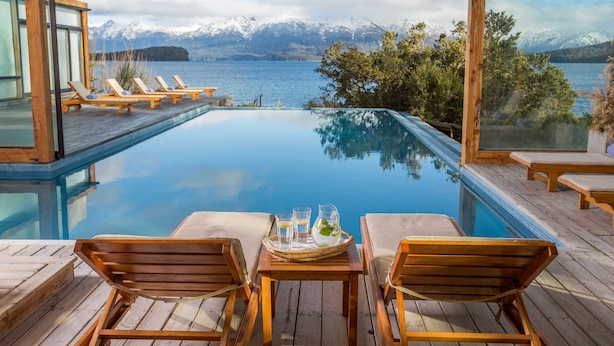 An infinity pool with wooden deck chairs and a table with drinks, overlooking a lake and mountains. - Correntoso Lake & River Hotel (Villa La Angostura)
