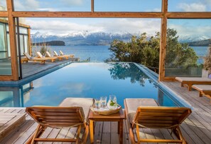Una piscina infinita con sillones de madera y una mesa con bebidas, con vistas a un lago y montañas.