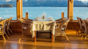 A dining table set for four with wicker chairs, a white tablecloth, and a view of a lake with mountains in the background. - Correntoso Lake & River Hotel (Villa La Angostura)
