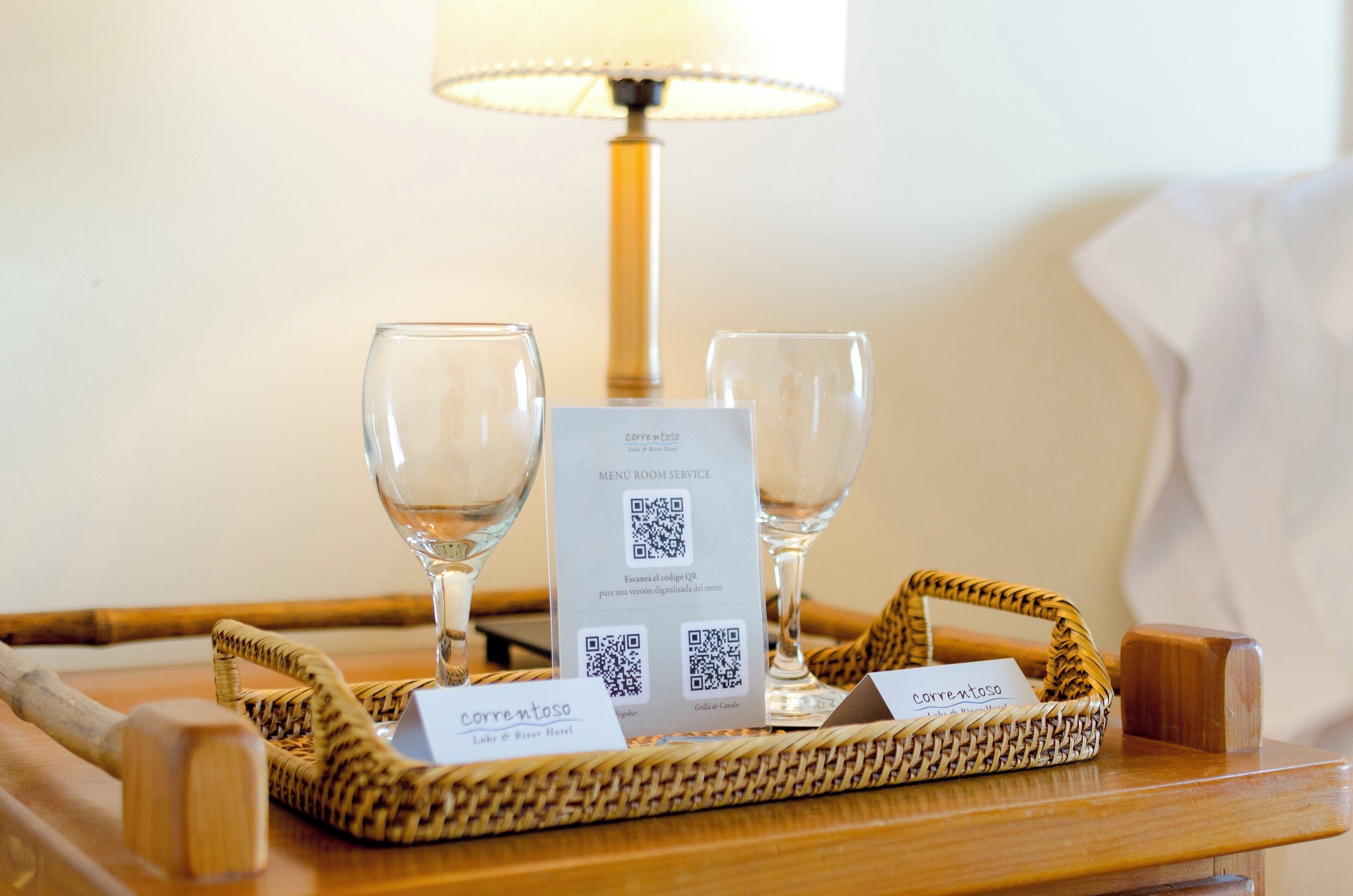 Uma bandeja com duas taças de vinho, um menu em código QR e uma etiqueta com nome sobre uma mesa de madeira.