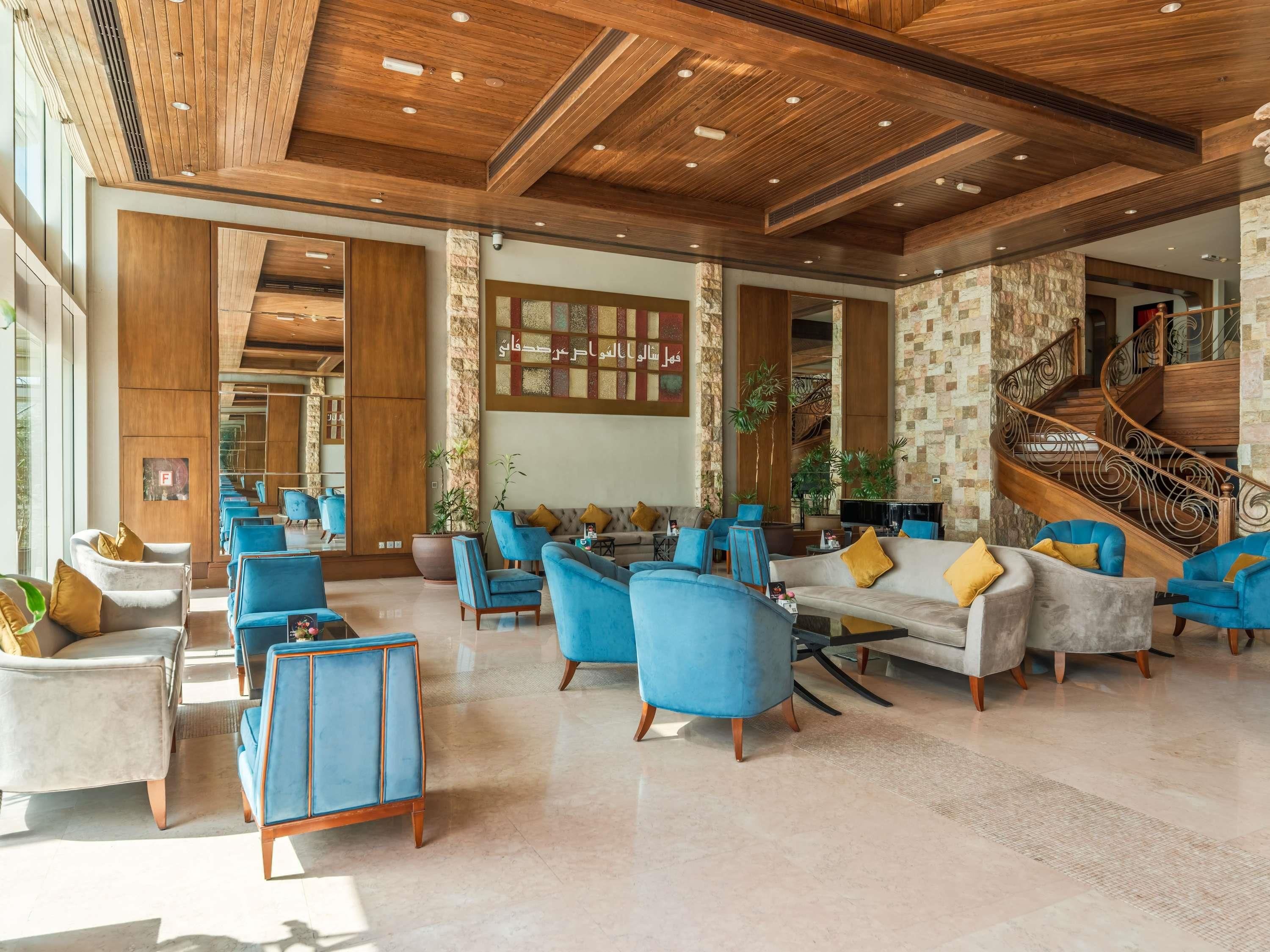 A spacious lobby with a high ceiling, wooden beams, and a stone accent wall. There are multiple seating areas with blue and white furniture, and a staircase with a decorative railing.