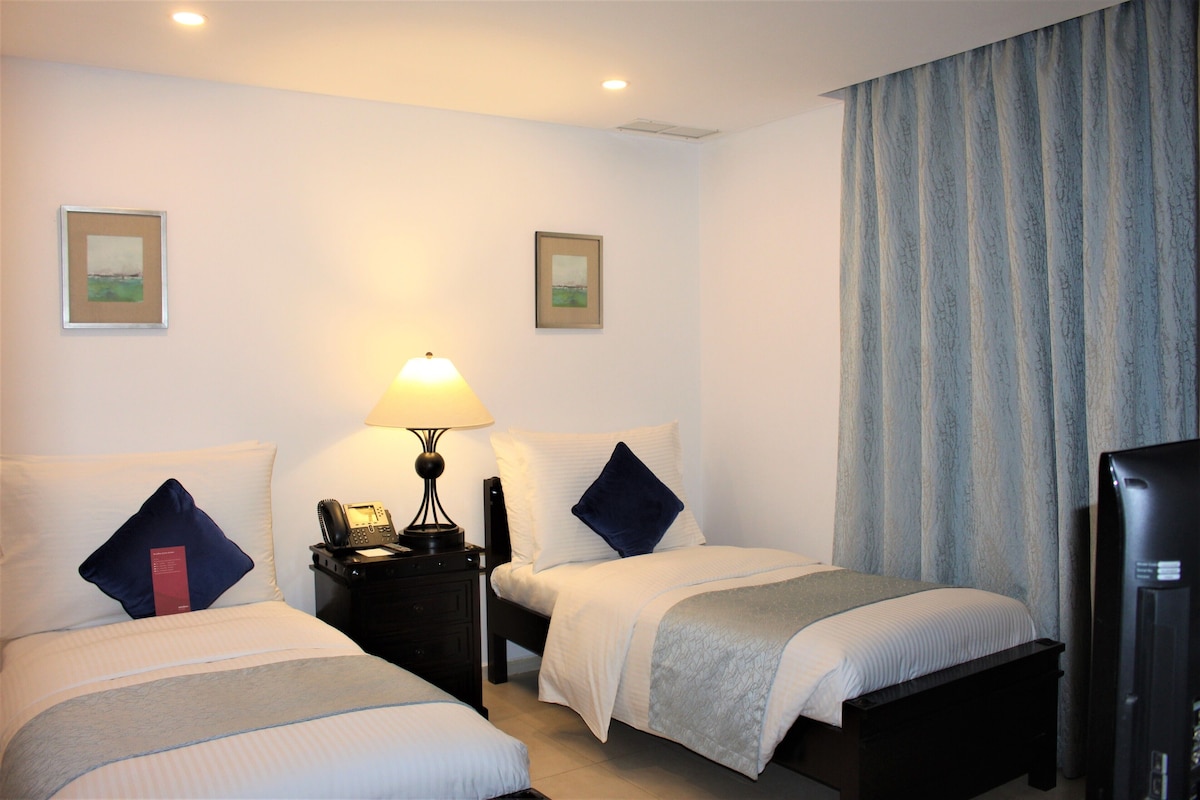 Una habitación de hotel con dos camas, una mesita de noche con lámpara, teléfono y televisión.