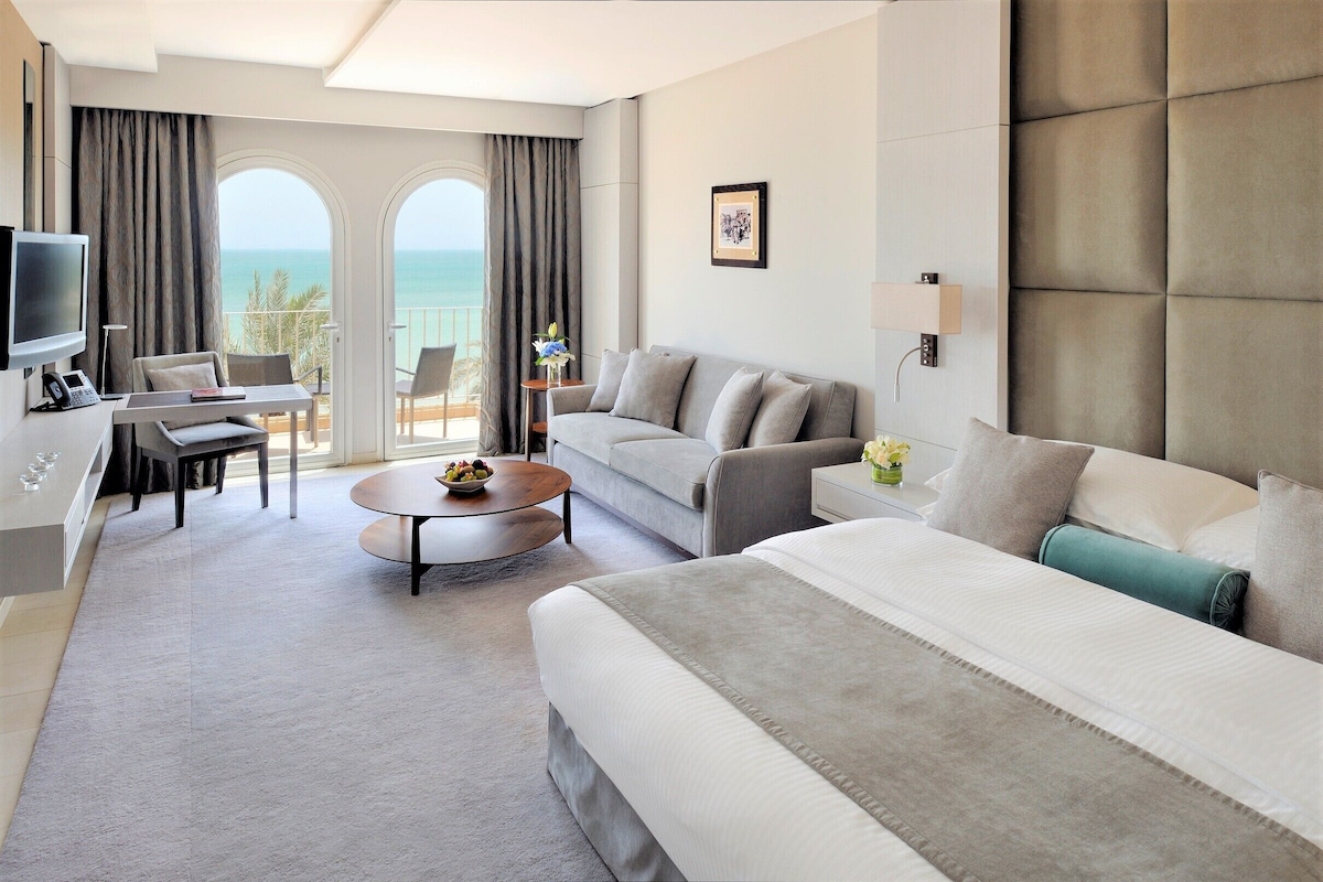 Habitación de hotel con cama, sofá, televisor, escritorio, silla, mesa de centro y vista al océano.